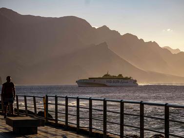 Gran Canaria, Spain: Ferry passing by mountains at sunset with a person strolling on the pier.