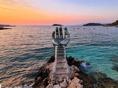 Ksamil, Albania. Sculpture of a hand emerging from the sea at sunset, with islands in the backdrop.
